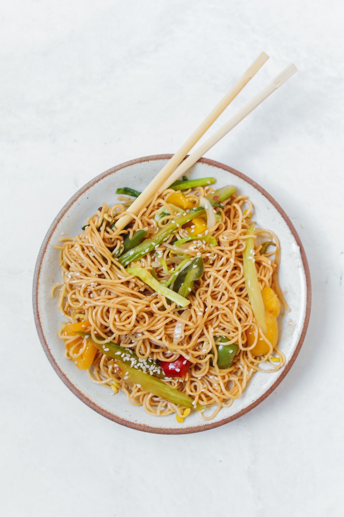 Chinese vegetable chow mein with thin noodles, scallions, peppers, sesame, and chopsticks.