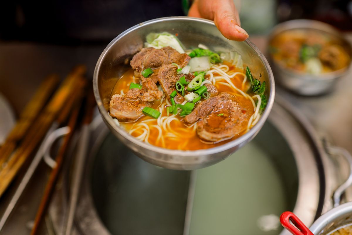 Braised beef noodle soup with beef chunks, scallions, greens, and orange broth in a metal bowl.